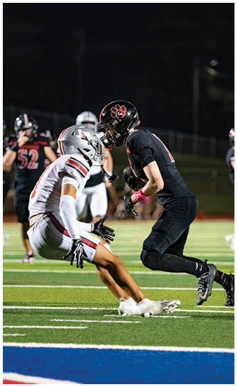 Two people in football uniforms are engaged in a play on a well-lit field at night with others in the background. Description generated by AI
