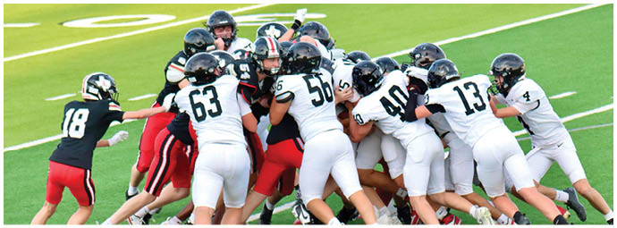 A group of people wearing football helmets and uniforms are engaged in a close physical play on a green field. Description generated by AI