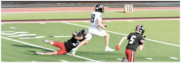 A person in a white uniform is running while another person in a black uniform is diving to tackle them and a third person in a black uniform is running nearby on a football field. Description generated by AI