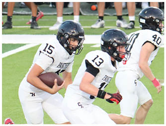 Three people in white football uniforms and black helmets are on a field, with one person holding a football and the others preparing to move. Description generated by AI