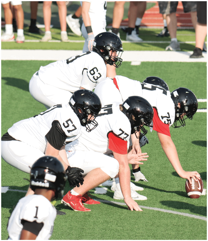 Several people wearing white football uniforms and black helmets are positioned on a football field, with some standing and others crouched in a ready stance. Description generated by AI