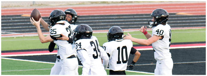 Four people in white football uniforms and helmets are on a field, with one person holding a football and others appearing to prepare for a play. Description generated by AI