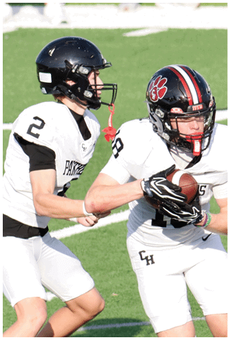 Two people in football uniforms and helmets are on a field, with one person holding a football and the other reaching toward them. Description generated by AI