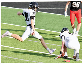 A person in a white football uniform is kicking a football held by another person in a white uniform on a green field while a third person in a black and red uniform stands nearby. Description generated by AI