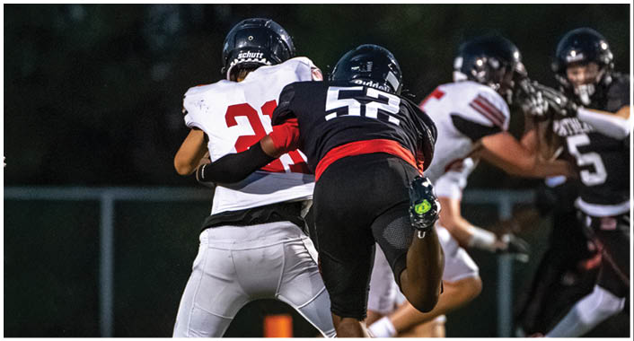 Two people wearing football helmets and uniforms are engaged in a play on a field with a dark background. Description generated by AI