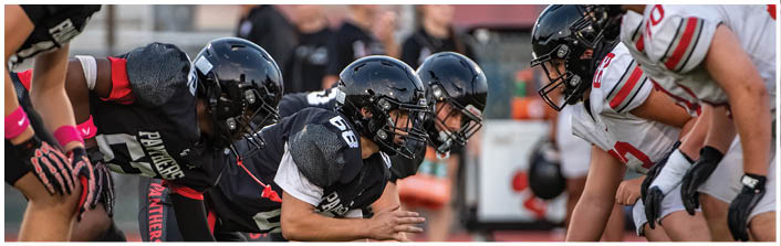 Four people in football uniforms and helmets are lined up in a ready stance on a field, preparing for a play. Description generated by AI
