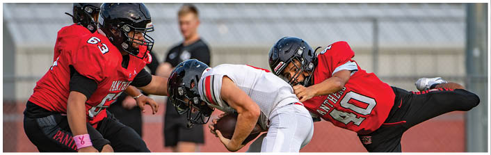Three people in football uniforms are engaged in a play on a field, with one person in white holding the ball and two others in red attempting to tackle. Description generated by AI