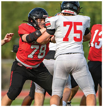 Two people wearing football helmets and white jerseys with red numbers are seen from behind on a field. Description generated by AI