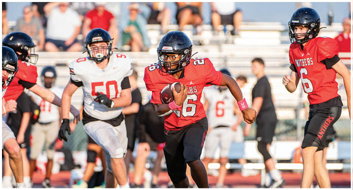 People wearing football uniforms are actively playing on a field with one person running while holding the ball and others chasing or preparing to block. Description generated by AI