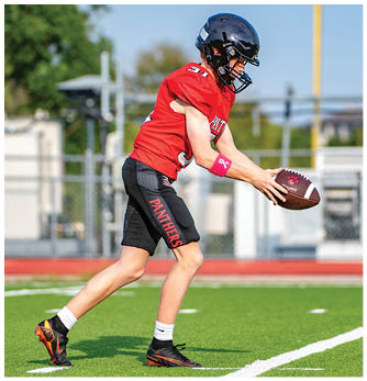 A person wearing a red sports jersey and black shorts is preparing to throw a football on a green field with a chain-link fence and trees in the background. Description generated by AI