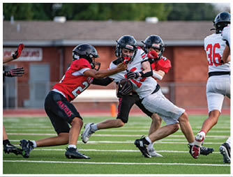 Two people in red sports uniforms are tackling a person in a white sports uniform who is holding a football on a green field. Description generated by AI