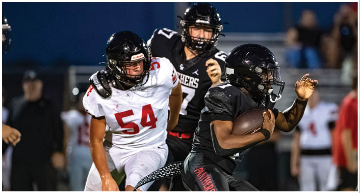 A person in black football gear is running with the ball while a person in white football gear with the number 54 is chasing them on a field. Description generated by AI