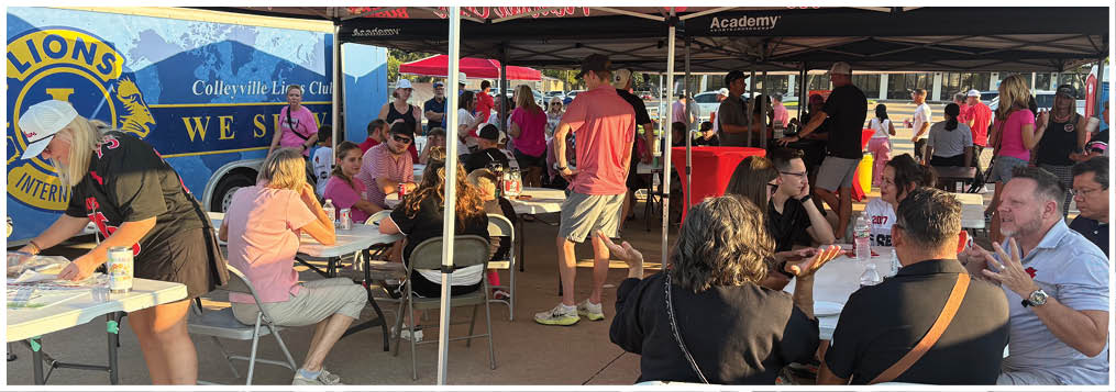 People are sitting and standing around tables under a tent at an outdoor event, engaging in conversation and activities. Description generated by AI