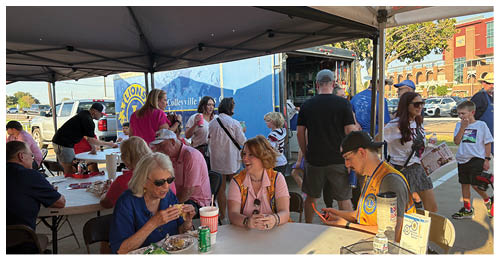 Several adults are sitting and standing around tables outdoors, eating, drinking, and engaging in conversation in a casual social setting. Description generated by AI