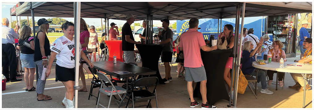 Adults and children are gathered under tents at an outdoor event with tables and chairs, some standing and talking while others sit and eat. Description generated by AI