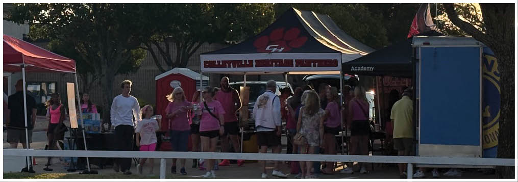 Adults wearing matching pink shirts are gathered under tents in an outdoor setting with trees and a sunset sky. Description generated by AI