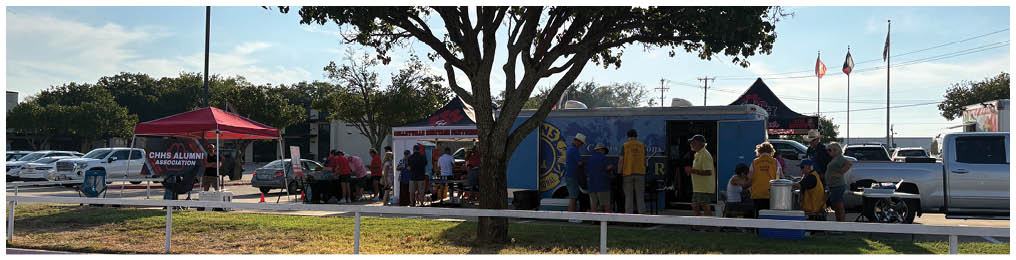 A group of people gathered under tents and around a food truck in a parking lot with a large tree casting shadows in the foreground. Description generated by AI