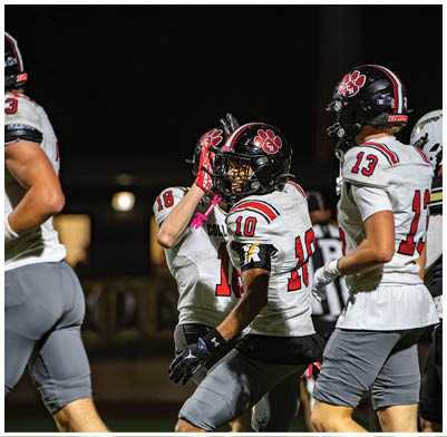 People wearing football uniforms are on a field at night, with one person in the center raising a hand to their helmet. Description generated by AI