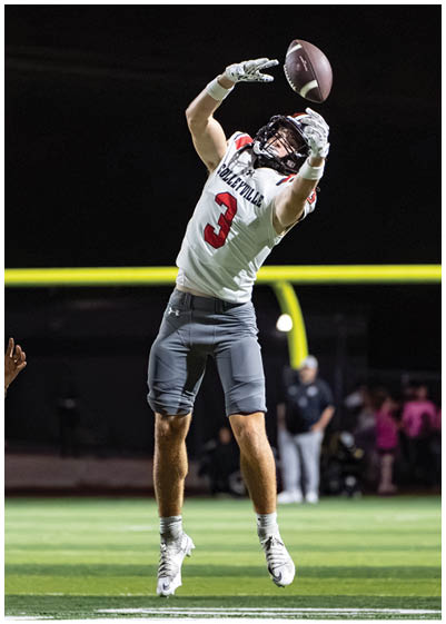 A person wearing a football uniform with the number three is jumping on a field at night, reaching up to catch a football near the goalposts. Description generated by AI