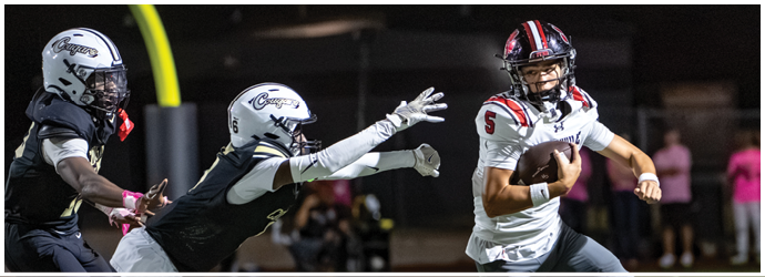 A person in a football uniform is holding a football while two other players in helmets and uniforms reach toward them on a nighttime field with a goalpost in the background. Description generated by AI