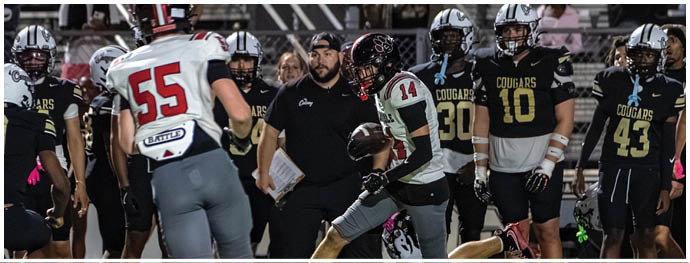A group of people in football uniforms are on a field with one person in a white and red uniform holding a football while others in black and yellow uniforms watch. Description generated by AI