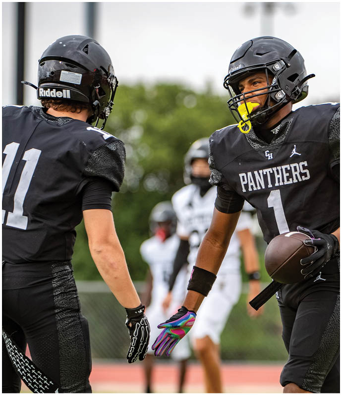 Two football players in black Panthers uniforms and helmets are reaching out to each other on a field with blurred players and greenery in the background. Description generated by AI