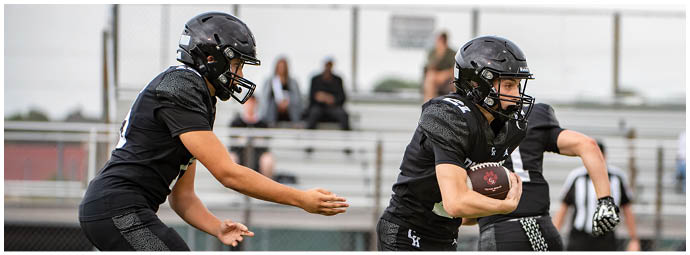 Two people wearing black football uniforms and helmets are actively engaged in a play on a football field with bleachers in the background. Description generated by AI