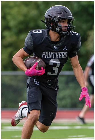 A person wearing a black football uniform with the number 3 and pink gloves is running on a field while holding a football. Description generated by AI