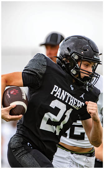 A person wearing a black football uniform with the word Panthers and the number 21 is running with a football while being pursued by others in white and black uniforms on a field. Description generated by AI
