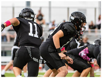 Two people in black football uniforms are actively playing on a green field with a referee and spectators in the background. Description generated by AI