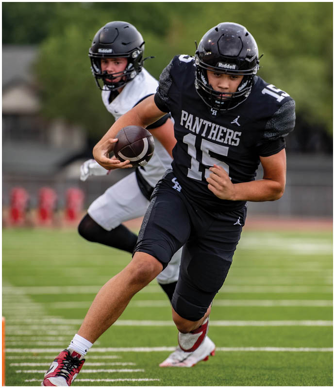 A person in a black football uniform with the word Panthers and number 15 is running with the ball while being chased by another person in a white uniform on a football field. Description generated by AI