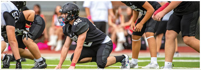 Two people in black football uniforms and helmets are positioned on a field with a blurred person in white in the background. Description generated by AI