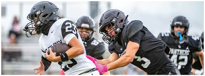 A group of people wearing football helmets and uniforms are actively engaged in a game, with one person holding the ball while others attempt to tackle or block. Description generated by AI