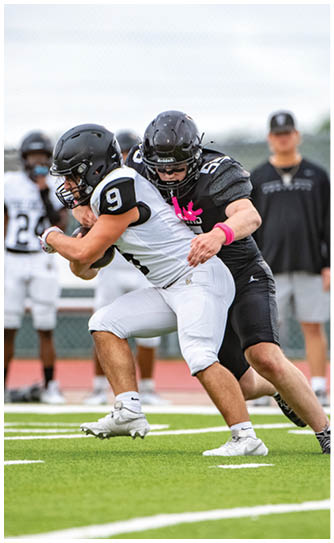 Two people in football uniforms are engaged in a tackle on a green field with others and a coach visible in the background. Description generated by AI