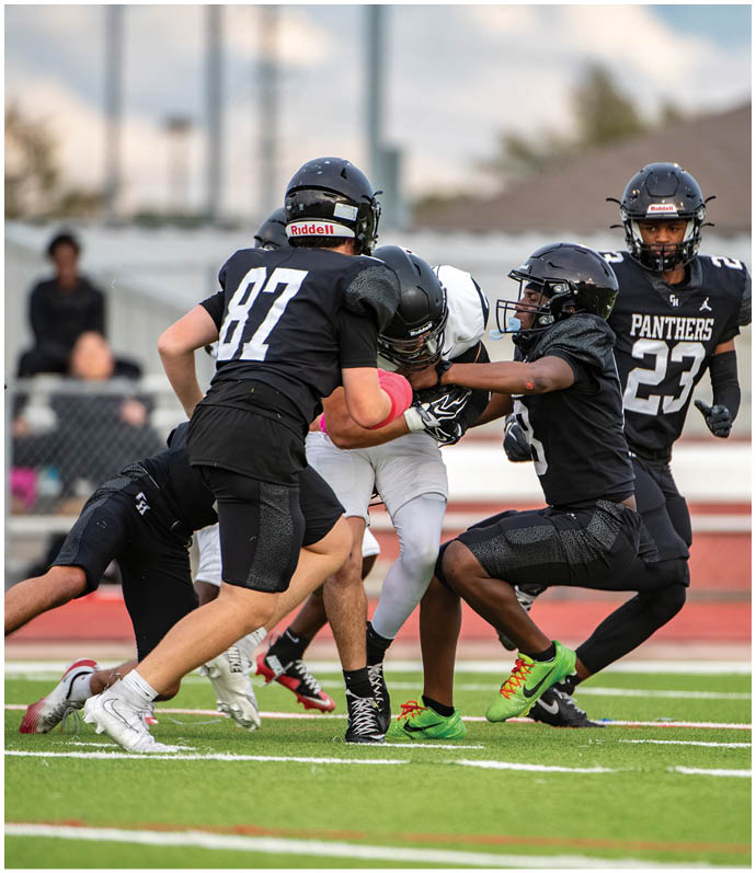 Several people in football uniforms are actively engaged in a tackle on a green field with a red running track in the background. Description generated by AI