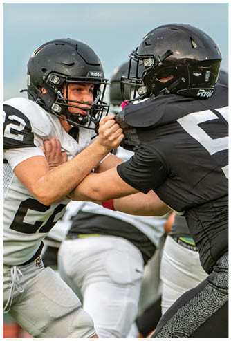 Two people wearing football helmets and uniforms are engaged in a blocking drill on a field with other players in the background. Description generated by AI