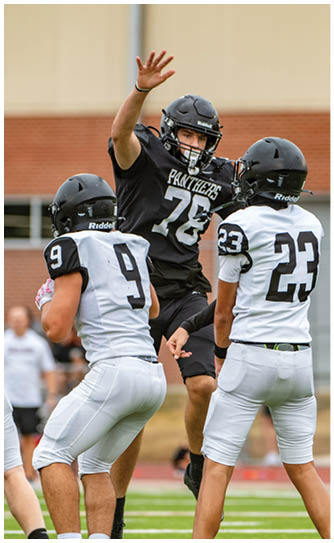 Three people wearing football helmets and uniforms are engaged in a play on a football field with a brick building and spectators in the background. Description generated by AI