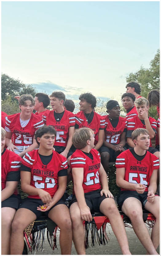 A group of people wearing red and black sports jerseys are sitting closely together on a decorated float outdoors with trees and a clear sky in the background. Description generated by AI