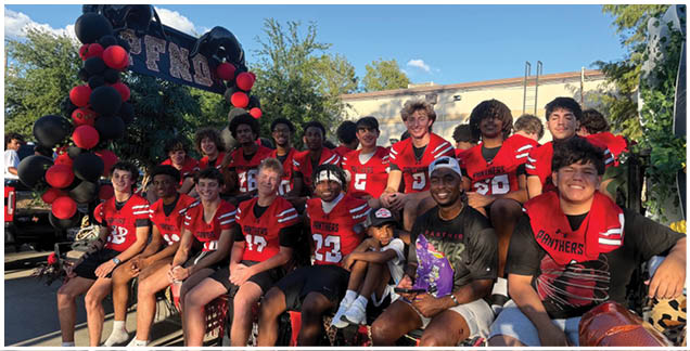 A group of people wearing red sports jerseys are smiling and posing closely together outdoors with trees and balloons in the background. Description generated by AI