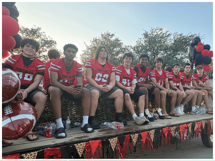 A group of people wearing red sports jerseys and shorts are sitting on a decorated flatbed trailer with football-themed decorations and balloons. Description generated by AI