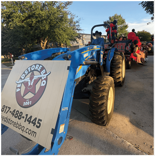 A person wearing a hat and sunglasses is sitting on a blue tractor decorated with red and black balloons in an outdoor setting with trees and a clear sky. Description generated by AI