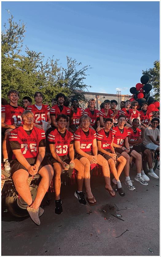 A group of people wearing red sports jerseys are sitting and standing together outdoors near a decorated float with red and black balloons. Description generated by AI
