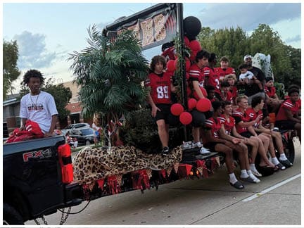 A group of people wearing matching red sports jerseys are sitting and standing on a decorated truck bed with balloons, a faux leopard print covering, and artificial palm trees, while one person in a white shirt stands near the truck's cab. Description generated by AI