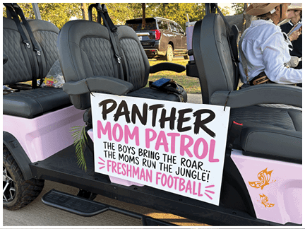 A sign with bold black and pink letters reads “Panther Mom Patrol" followed by the phrases "The boys bring the roar, the moms run the jungle" and "Freshman" placed on a seat inside a vehicle. Description generated by AI