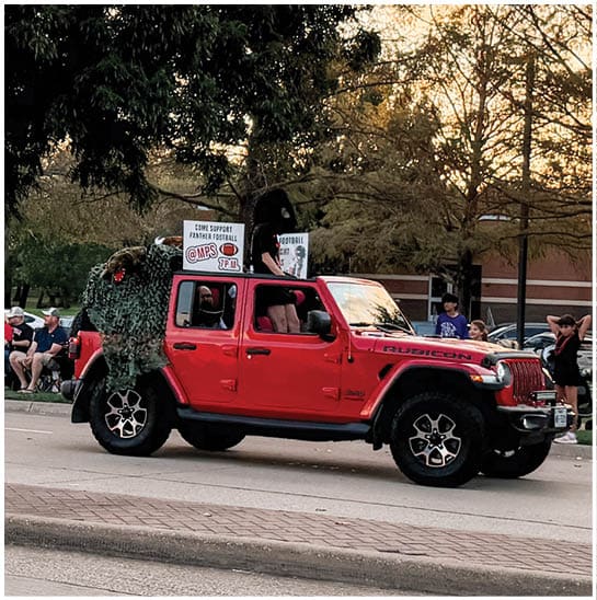 A red Jeep Rubicon is parked on a street with several people standing and sitting nearby, and trees and a building are visible in the background. Description generated by AI
