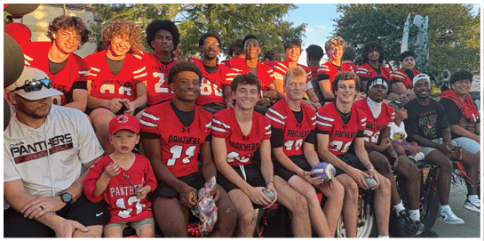 A group of people wearing red and black sports jerseys and casual clothing are sitting together outdoors on a bench with balloons and trees in the background. Description generated by AI
