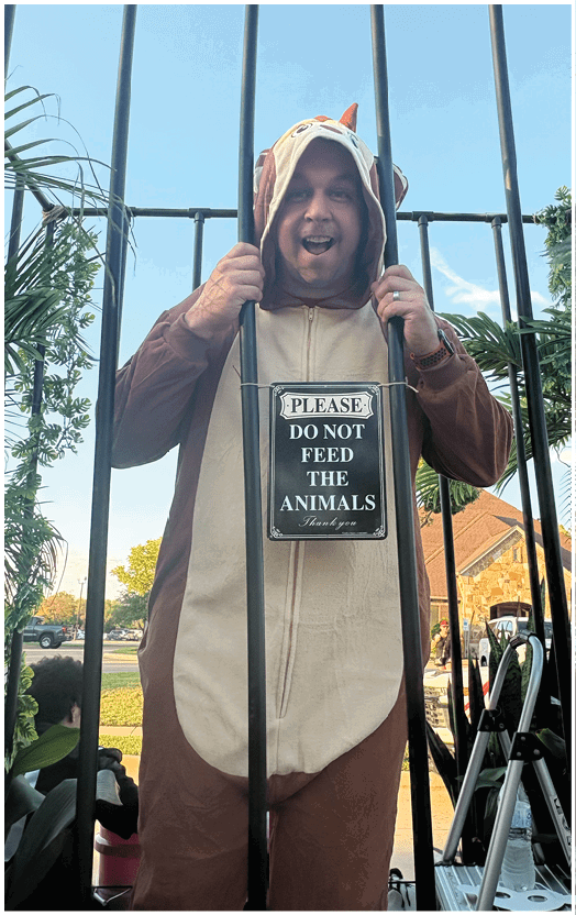 A person wearing a brown and beige animal onesie is standing behind a metal fence with a sign that reads “PLEASE DO NOT FEED THE ANIMALS." Description generated by AI