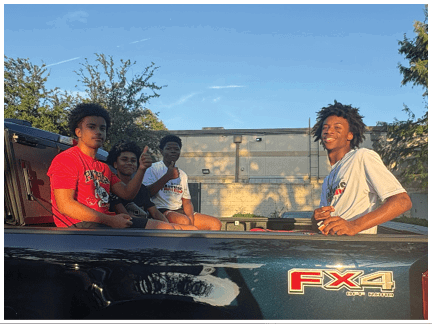 Four people with casual clothing and natural hairstyles are sitting and smiling in the back of a black pickup truck with an FX4 Off Road decal, parked outdoors near a building and trees in the late afternoon sunlight. Description generated by AI