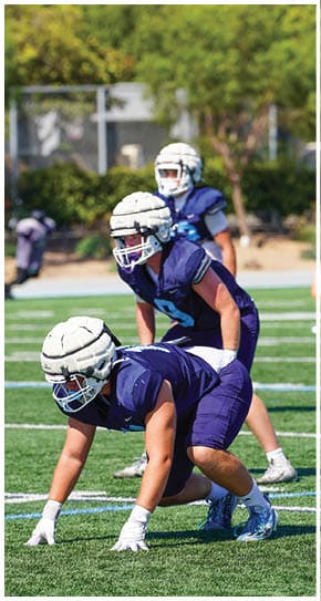 Three football players in blue and white uniforms running on a field. Description generated by AI