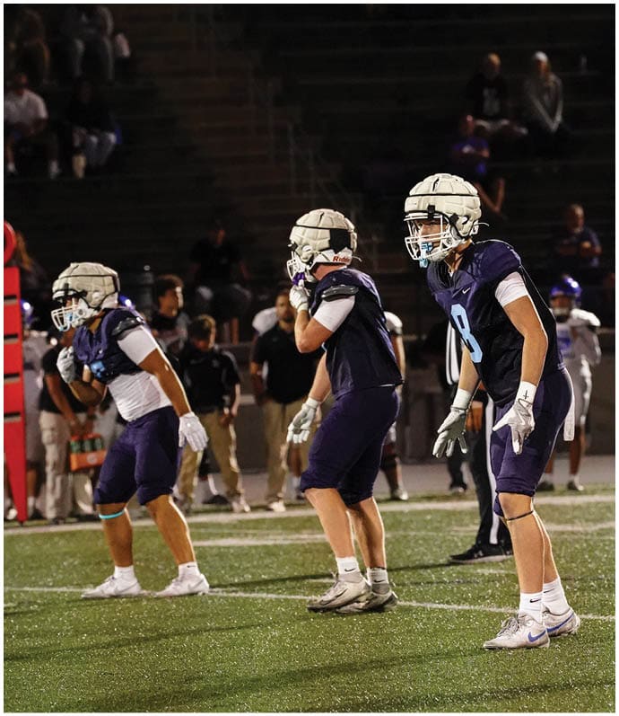 Three football players in blue and white uniforms running on a field. Description generated by AI